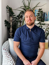 Ross Chaplain sitting on a chair in front of a selection of house plants and books, wearing a blue t-shirt and smiling at the camera.