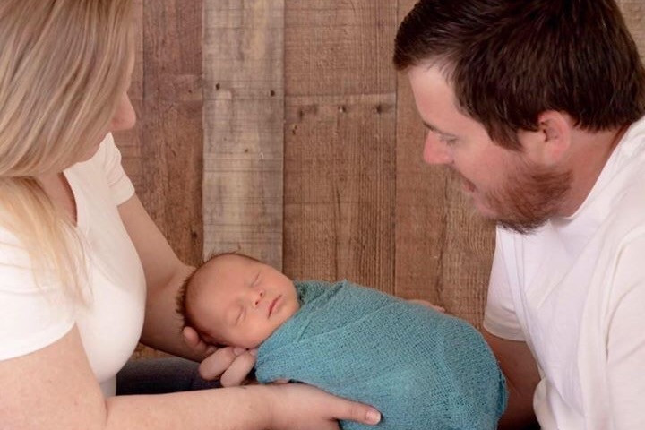 A couple holding a tiny, sleeping baby with brown hair who is wrapped in a blue blanket.