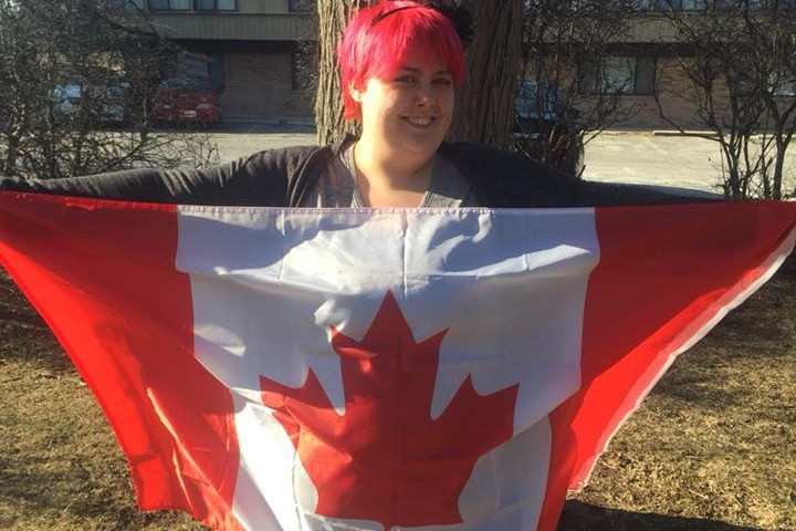 A photo of a person with short, hot pink/reddish hair holding a Canadian Flag. They are light-skinned and smiling.