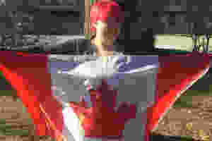 A photo of a person with short, hot pink/reddish hair holding a Canadian Flag. They are light-skinned and smiling.