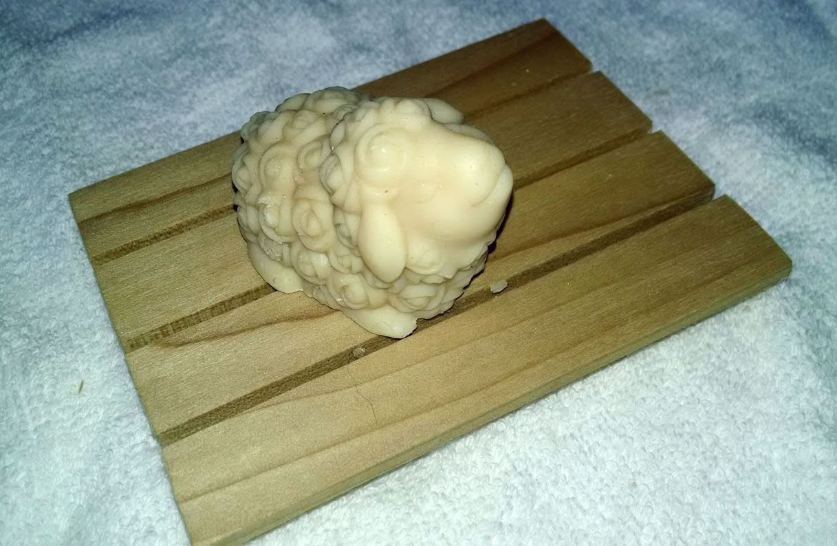 A small sheep or angora goat sculpture, resting on an oak wooden soap dish.