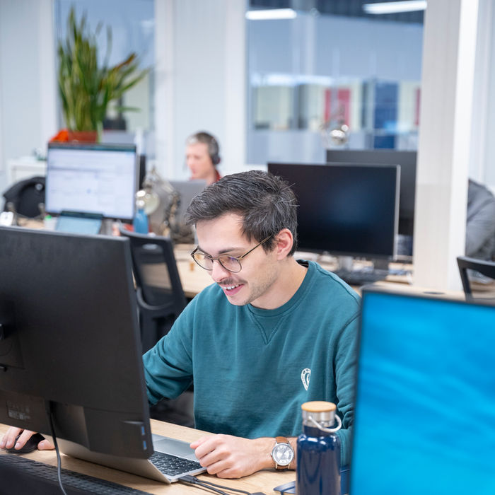 Homme souriant travaille sur ordinateur avec un collègue en arrière-plan dans un bureau.