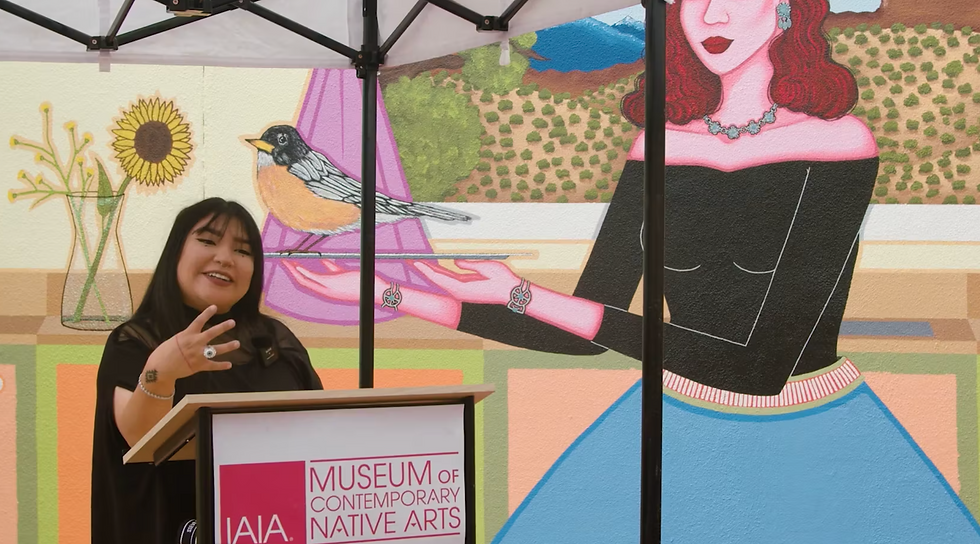 Color photo of artist Robyn Tsinnajinnie speaking while in front of an elargement of one of her paintings