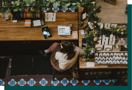 Aerial perspective of a retailer sitting on a stool at the cashwrap with an iPad showing the point-of-sale screen. The shop owner is surrounded by merchandise including spools of ribbon and gift tags with different initials.