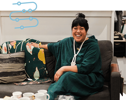 Chris who sits casually on a love seat inside of a Women's Apparel and Lifestyle store. Her hair is in a bun, her arm is draped along the back of the sofa, and she shares a knowing laugh.