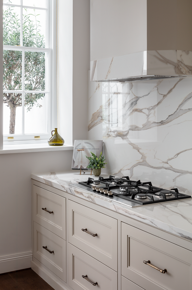 Close-up of a modern gas hob set into a polished marble worktop with matching splashback, surrounded by cream shaker-style cabinetry and natural light from a nearby window.