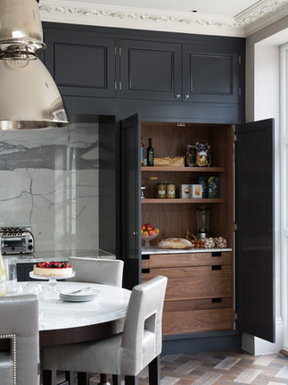 Luxury kitchen with dark cabinetry, open wooden larder, marble splashback, and a round dining table set for guests.