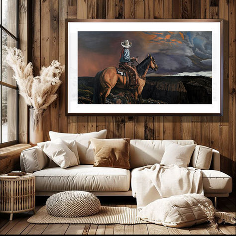Oil painting of a lone rider on the plains with a stormy sky by Michael Steward, in a mahogany frame, displayed in a rustic room with a wooden wall, white canvas sofa, and raw wooden floorboards.