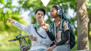 Two people sitting outdoors under trees, holding coffee cups. One points, while the other listens with headphones. A bike is nearby.
