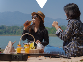 Two women enjoy a picnic by a lake with mountains in the background. One drinks a beverage, surrounded by fruit, juice, and a guitar.