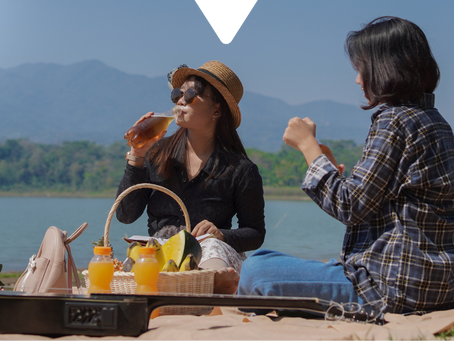 Two women enjoy a picnic by a lake with mountains in the background. One drinks a beverage, surrounded by fruit, juice, and a guitar.
