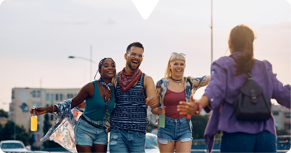 Four friends joyfully greet each other outdoors. Two women and a man face a person in purple, with a warm, sunny cityscape behind them.