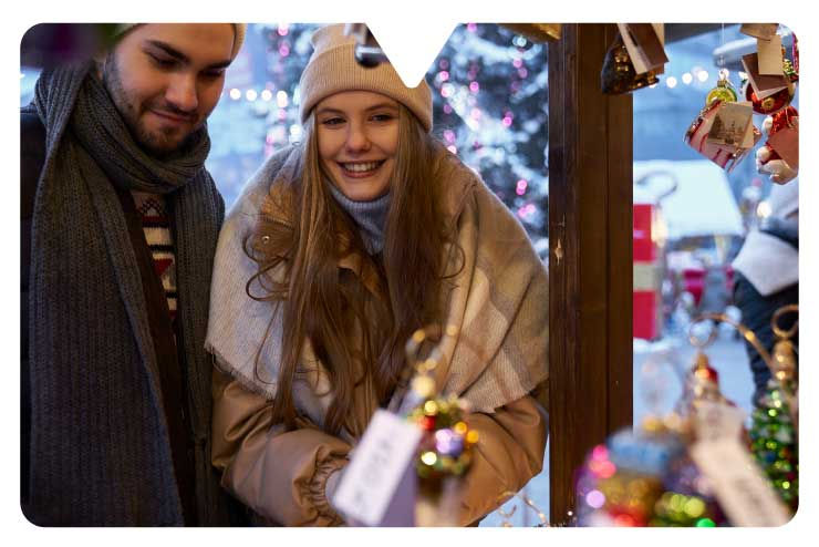 Two people in winter clothing smiling at a holiday market stall with colorful ornaments. Festive lights and snow in the background.