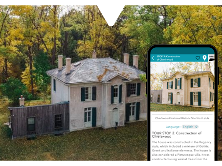 Two-story house in autumn forest, beige with green shutters. Nearby phone screen displaying the Driftscape digital tourism app shows tour info about a particular tour stop. Historic vibe.