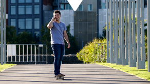 Man skateboarding on a paved path, holding a phone and smiling. Modern buildings and greenery line the sunny, open area.
