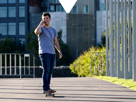 Man skateboarding on a paved path, holding a phone and smiling. Modern buildings and greenery line the sunny, open area.