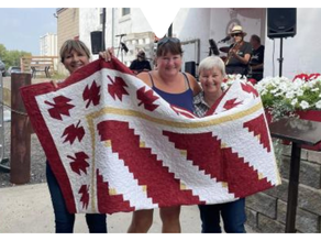 Three smiling women hold a red and white quilt with leaf patterns. Musicians play in the background; flowers and a building are visible.