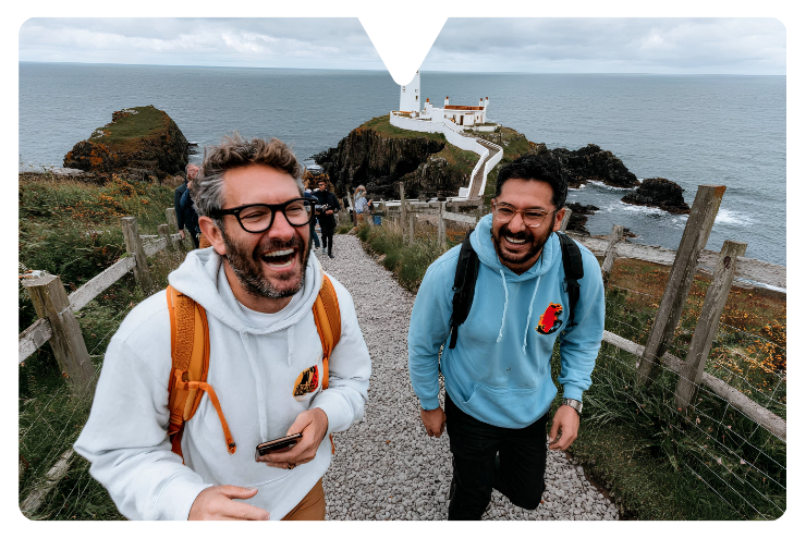 Two men in hoodies laughing on a coastal path, with a lighthouse and ocean in the background. The mood is joyful and scenic.