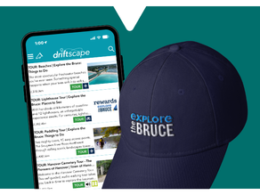 Phone displaying the Driftscape digital tourism app showing various tours and images. A navy cap with "Explore the Bruce" logo is in foreground on teal background.