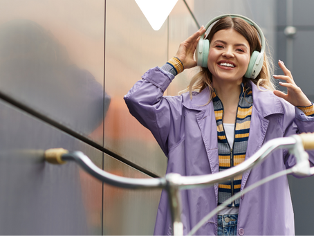Woman in a purple coat and headphones smiles while touching her hair. Bicycle handlebars in foreground, modern building in background.
