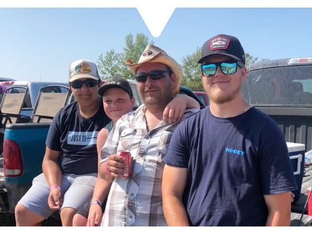 Four people sitting on a truck tailgate wearing hats and sunglasses. One holds a soda can. Clear sky and parked cars in the background.