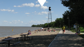 People enjoy a sunny day on a sandy beach beside a lake. A tall water tower is visible in the background. Lush trees line a walkway.