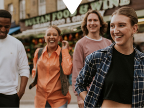 Four people smiling and walking outdoors by a food and wine store. One wears a checkered shirt. Joyful mood, casual attire.