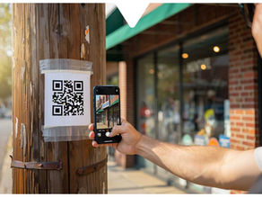 Person scans a QR code taped to a wooden pole with a smartphone. Background shows a storefront with a green awning on a sunny day.