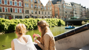 Two women sit by a canal eating ice cream. A duck swims by, while people paddleboat nearby. Historic buildings line the sunny background.