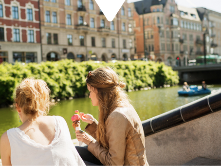 Two women sit by a canal eating ice cream. A duck swims by, while people paddleboat nearby. Historic buildings line the sunny background.