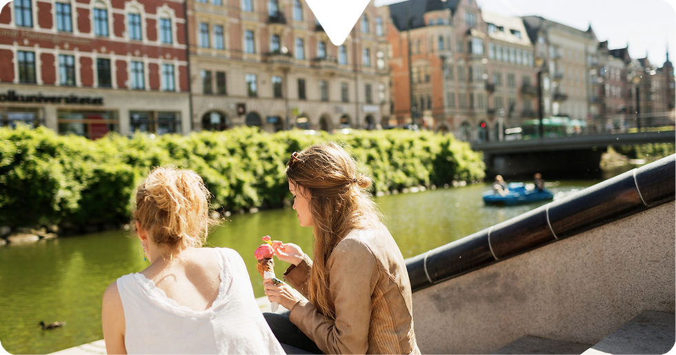 Two women sit by a canal eating ice cream. A duck swims by, while people paddleboat nearby. Historic buildings line the sunny background.