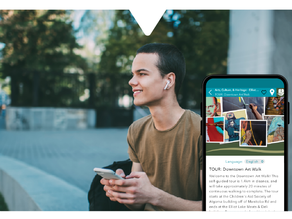 A person sits outdoors smiling with earbuds in, holding a smartphone. The phone screen shows a Downtown Art Walk tour via the Driftscape digital tourism app. Background is blurred trees.