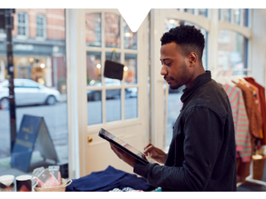 Man using a tablet with an ai business directory via the Driftscape app, standing near a window. Outside, cars and buildings are visible. Casual, focused atmosphere.