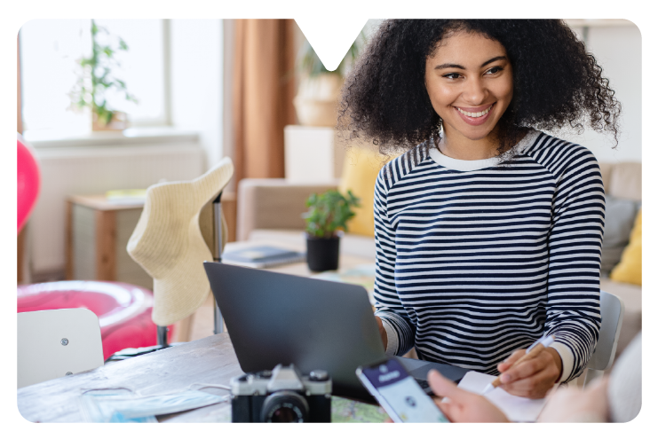 Woman in a striped shirt smiles while using a laptop at a table with a camera and plant. Bright indoor setting, joyful mood.