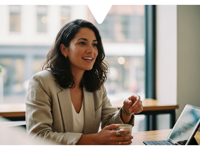 Woman in a beige blazer smiling and talking, holding a cup near a laptop displaying the Driftscape digital tourism app. Bright office setting with large windows in the background.