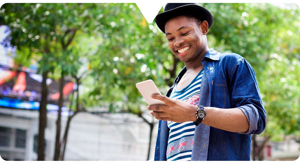 Smiling person in a hat, using a smartphone outdoors. Wearing a striped shirt and denim jacket. Green trees in the background. Bright mood.
