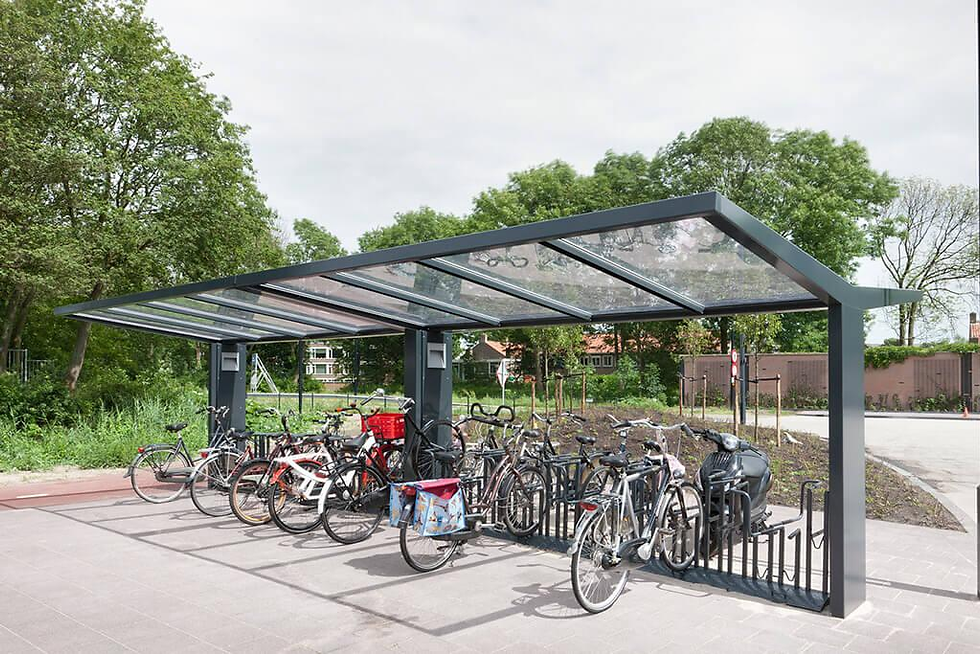 Covered bike parking shelter with many bicycles parked under a glass roof, surrounded by trees on a quiet street.