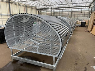 Row of metal mesh and corrugated structures inside a greenhouse. Brown floor, transparent ceiling, and light streaming through. No text visible.