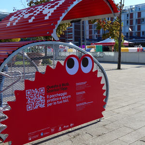 Red bicycle rack with cartoon eyes and Italian text promoting a bike parking app in a sunny urban plaza, with trees and a modern building.