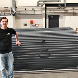 Man in a black shirt standing next to a Fietshangar 2.5 bike storage in a workshop. The setting is industrial, with pipes and pallets.