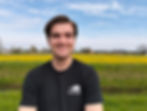 Man smiling in a black t-shirt with Fietshangar logo, standing in a field of yellow flowers under a blue, partly cloudy sky.