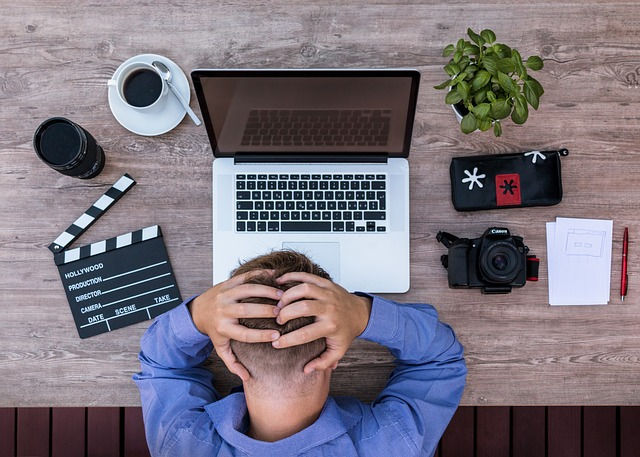 Homem com as mãos na cabeça em frente a uma mesa com notebook, câmera fotográfica, xícara, claquete e papel