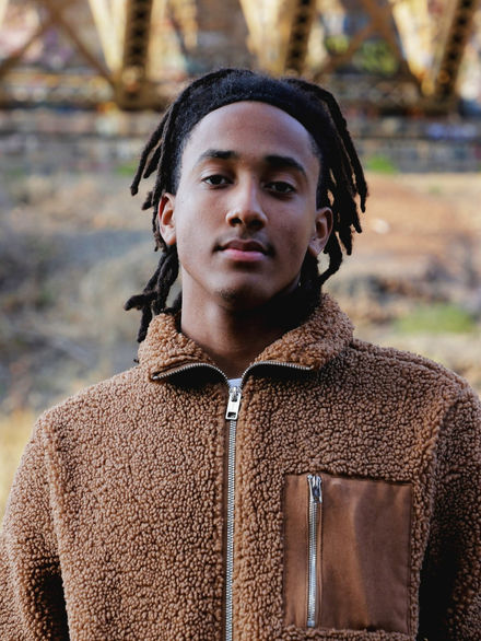A boy in a fuzzy brown jacket standing outside with his hands in his pocket