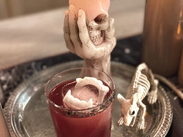 A Dracula's Kiss cocktail sitting on a silver tray in front of a skull candle and next to a rat skeleton for Halloween.