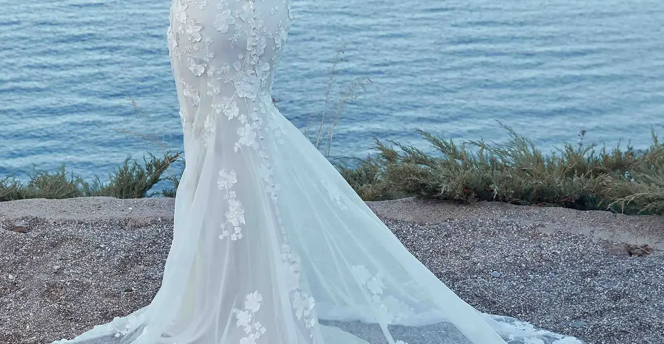 Back of Eddy K strapless fit-n-flare wedding gown with lace over tulle and an unlined train on bridal model standing near the beach.