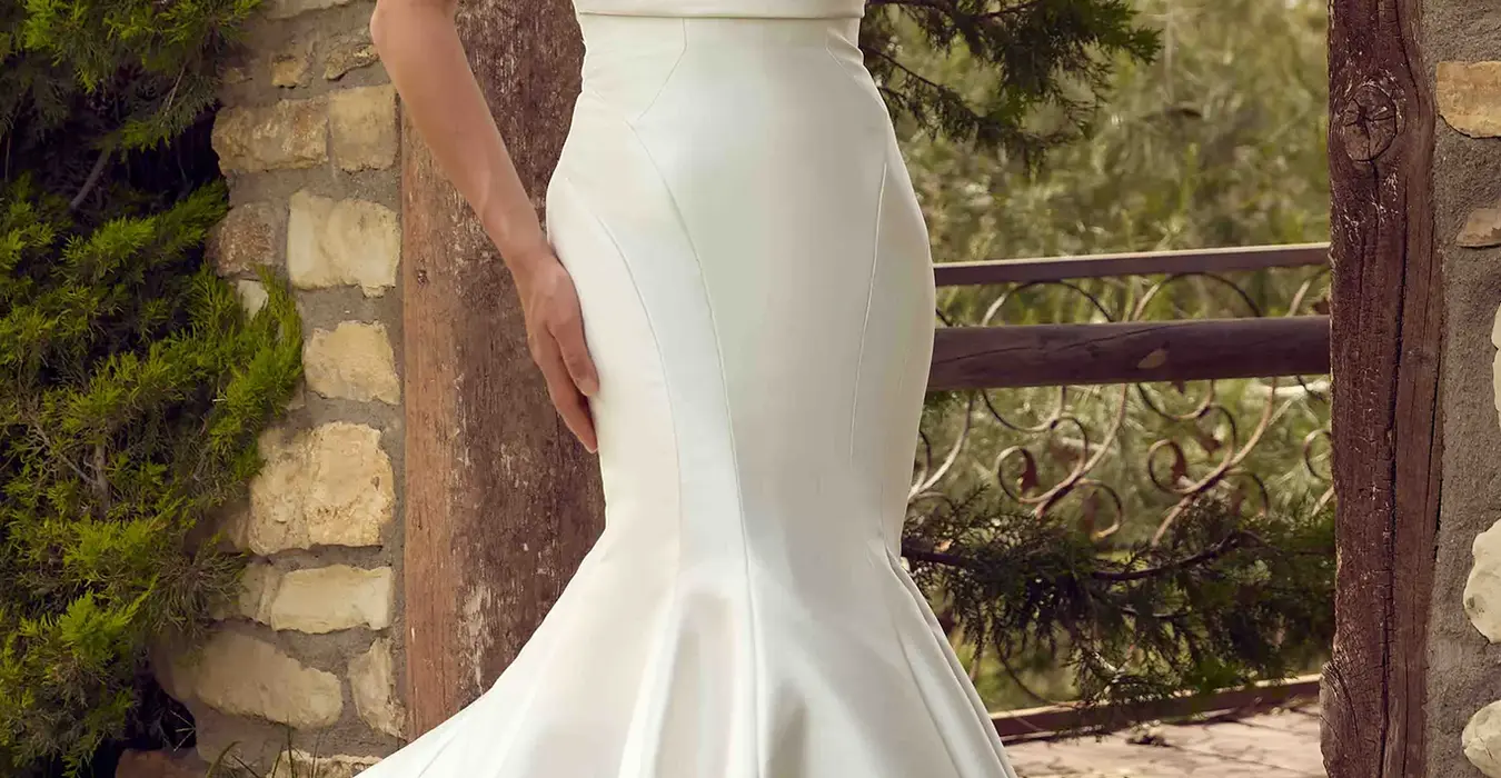 Eddy K mikado-satin mermaid wedding dress with a sweetheart neckline and a strapless body on a bridal model standing on a sunny grass field filled with trees.