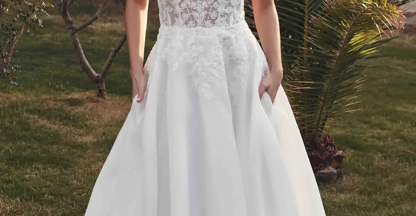 Eddy K sleeveless ballgown wedding dress with beaded ivory lace unlined bodice and organza skirt with pockets on bridal model standing in greenery garden.