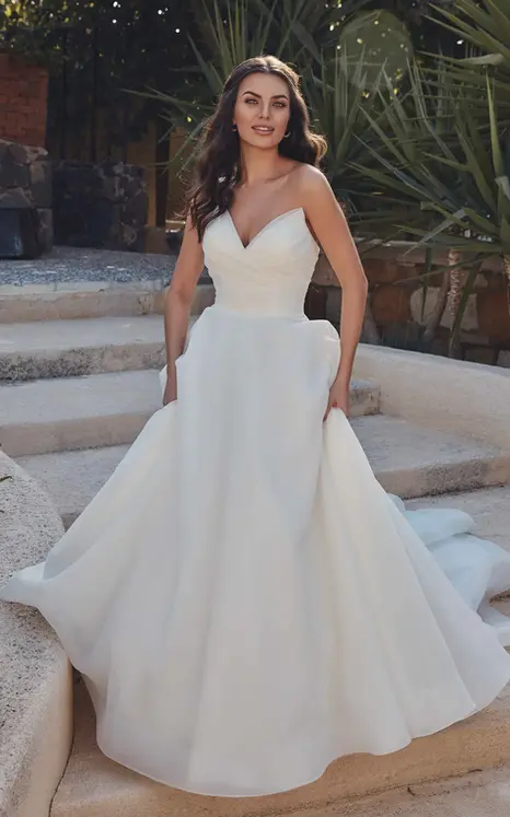 Eddy K organza ball gown in ivory with cat eye neckline on bridal model standing on stone stairs with greenery in background holding the skirt of her dress.