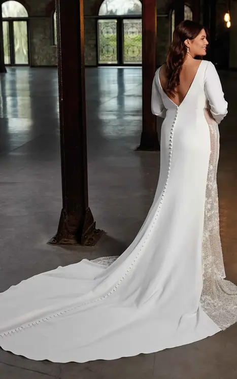 Back of Eddy K ivory crepe long sleeve wedding dress with side panels and buttons to end of train on bride standing in warehouse.
