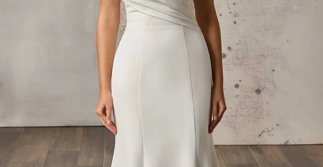 Eddy K crêpe fit-n-flare wedding dress with a sweetheart neckline and an off-shoulder body on a bridal model standing in a bridal studio.